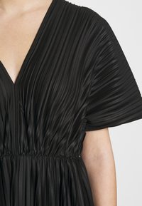 Gros plan d'une personne portant une robe noire plissée à manches courtes et taille froncée.