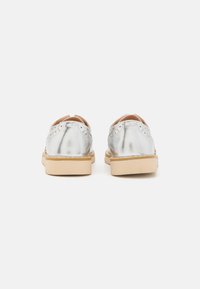 Scarpe metalliche argento con dettagli brogue, punta tondeggiante e suola in gomma beige. Presentano una texture liscia e un tacco basso.