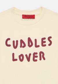 Tricou de culoare crem cu text roșu „CUDDLES LOVER” și o etichetă roșie MAX&Co. în interiorul gulerului.