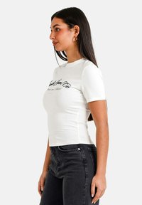 Hvid tætsiddende t-shirt med korte ærmer og rund halsudskæring, prydet med sort tekstdesign på forsiden, parret med mørke jeans.