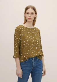 Olivengrübluse mit kleinen weißen und rosa Blumenmustern, Rundhalsausschnitt, dreiviertelÄrmel und lockerer Passform. Kombiniert mit blauen Jeans.