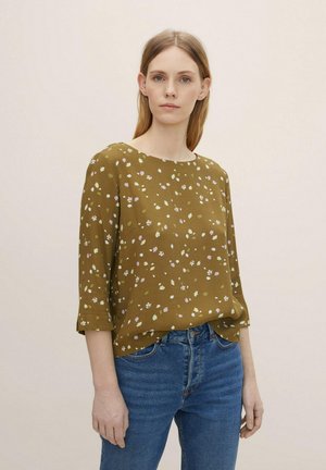 Olivengrübluse mit kleinen weißen und rosa Blumenmustern, Rundhalsausschnitt, dreiviertelÄrmel und lockerer Passform. Kombiniert mit blauen Jeans.