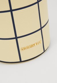 Superficie in pelle beige con motivo a griglia blu navy e lettere in metallo dorato che scrivono "COCCINELLE" sul bordo inferiore.