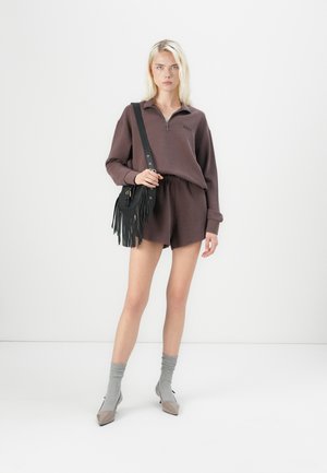 Bruine zip-up sweatshirt met ribbelstructuur, gecombineerd met bijpassende bruine shorts. Model houdt een zwarte franje schoudertas vast. Grijze sokken en puntige hakken.