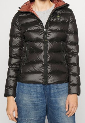 Femme portant une doudoune matelassée noire avec une capuche couleur rouille, un t-shirt gris et un jean bleu, debout les bras détendus.