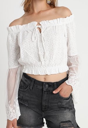 Biały top typu crop z wyciętymi ramionami i oczkami, z przezroczystymi koronkowymi rękawami w kształcie dzwonków, zestawiony z czarnymi, poszarpanymi szortami jeansowymi z wysokim stanem.