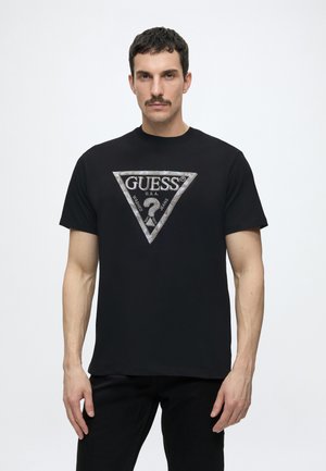 Homme aux cheveux foncés et moustache portant un T-shirt noir avec un grand triangle argenté du logo Guess centré sur la poitrine.