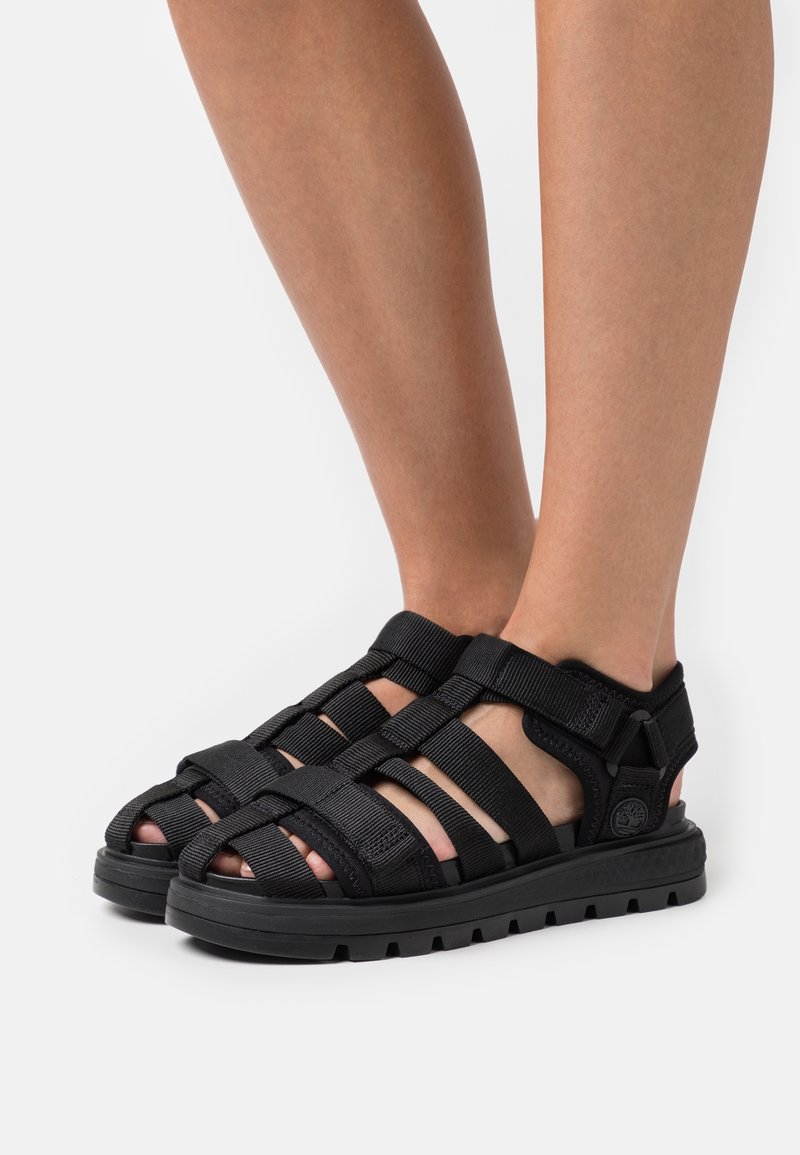 Personne portant des sandales noires à brides avec des semelles épaisses, debout sur une surface blanche, montrant les vues latérale et avant des chaussures.
