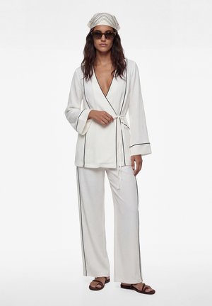 Mujer con conjunto de ropa de descanso blanca con ribetes negros, pañuelo en la cabeza, gafas de sol y sandalias marrones, posando frente a un fondo blanco.