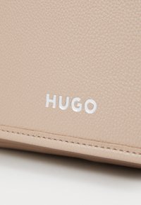 Lys beige teksturert lær med preget "HUGO"-logo i hvitt. Synlig søm langs kantene forbedrer holdbarheten.