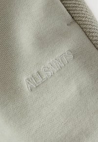 Vaaleanharmaassa kankaassa on pehmeä, sileä pinta, jossa on brodeerattu "ALLSAINTS"-logo hiukan tummemmalla langalla.