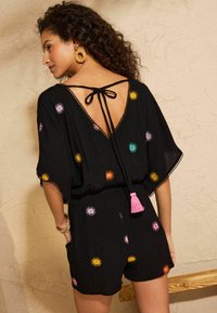 Schwarzer Jumpsuit mit kurzen Ärmeln, verziert mit mehrfarbiger Blumenstickerei, Rückenverschluss mit Bindeband und pinkfarbener Troddeldetail am Taillenbereich.