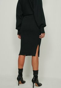 Nainen, jolla on päällään musta ribbiraitainen midi-hame, jossa on sivuhaalari, musta pitkähihaisa paita ja mustat korkeat nilkkasaappaat, seisoo.