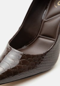 Chaussure à talon haut brun foncé à bout pointu en cuir synthétique texturé effet crocodile brillant avec semelle intérieure rembourrée.