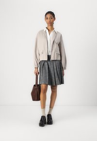 Lys beige cropped jakke med elastiske manchetter over en hvid skjorte med knapper. Plisseret grå nederdel, tan sokker, sorte platform loafers og en brun taske.