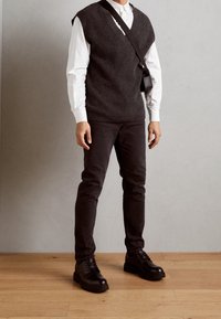 Chaleco de punto gris oscuro sobre una camisa blanca con cuello, combinado con pantalones oscuros y zapatos negros con cordones brillantes. Incluye un pequeño bolso negro de hombro.