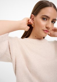 Helles Rosa, Pullover mit Rundhalsausschnitt und strukturiertem Strick, kombiniert mit goldenen Creolen. Die Person passt die Ohrringe nahe ihrem Ohr an.