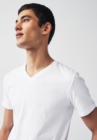 T-shirt bianca in cotone con scollo a V, maniche corte, tessuto liscio e vestibilità comoda, indossata da una persona che guarda verso l'alto su sfondo neutro.