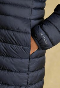 Tumes värvi puffjakki horisontaalsete õmblustega, küljetasku ja brändinime "JOULES" hõbeda aktsendiga mansetil.