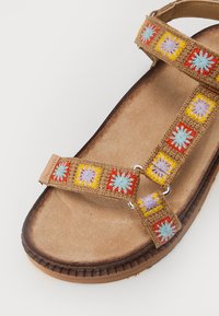 Sandale beige avec une sangle tissée ornée de motifs floraux colorés en rouge, jaune et bleu, et une semelle intérieure texturée et profilée.