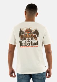 Camiseta de algodón color beige claro con un colorido gráfico de una furgoneta aparcada, árboles y sillas de campamento, con el texto "Timberland Rugged Active Gear" debajo.