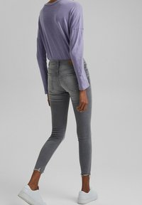 Top de manga larga color lila claro combinado con jeans ajustados grises que tienen dobladillo desgastado y bolsillos traseros, completado con zapatillas blancas.