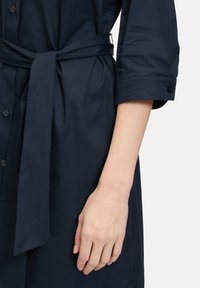 Abito blu navy con abbottonatura e maniche arrotolate, caratterizzato da una cintura annodata in vita. Tessuto morbido con una leggera lucentezza.