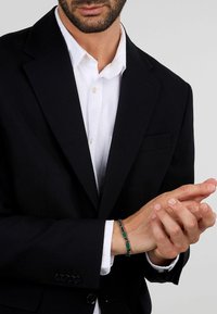 Blazer nero sopra una camicia bianca, con un braccialetto verde di perline al polso. Focus sulla stoffa del completo e sulla texture e design del braccialetto.