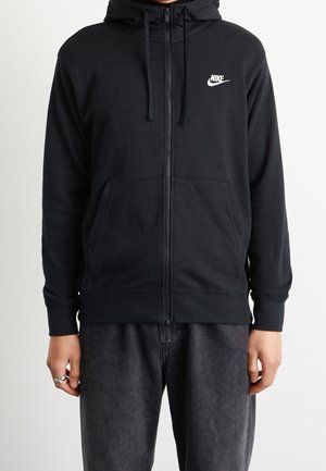 Personne portant un sweat à capuche Nike noir zippé avec poches avant et cordon de serrage à la capuche, associé à un pantalon gris foncé, debout devant un fond blanc.