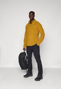 Gele fleecejas met ritssluiting, zwarte broek en laarzen. Rugzak over één schouder gehangen. Model staat tegen een effen witte achtergrond.