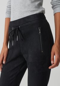 Jogging noir en suède avec une taille à cordon, deux poches avant et une poche latérale zippée. Texture lisse et coupe ajustée.