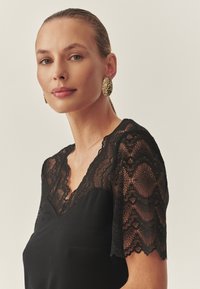 Črna obleka z kratkimi čipkastimi rokavi, izrezom v obliki črke V in gladko teksturo. Zlati ovalni uhani dodajajo okrasni poudarek.