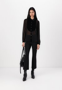 Camisa preta transparente com mangas longas e colarinho de veludo, com botões na frente e punhos ajustados, combinada com calças pretas de boca de sino e botas de tornozelo com bico apontado.
