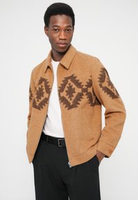 Fleecejakke i varm brun med diamantmønster i mørkebrun. Har lynlåslukning foran, krave og sidelommer.