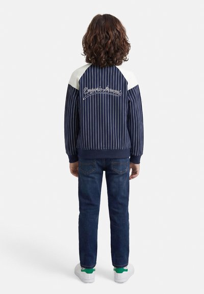 Παιδί με καστανά σγουρά μαλλιά που φοράει σκούρο μπλε σακάκι με ρίγες, σκούρο μπλε τζιν και λευκά αθλητικά παπούτσια με πράσινες λεπτομέρειες, στέκεται με την πλάτη γυρισμένη.