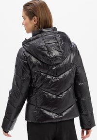 Femme portant une doudoune noire brillante avec capuche, de dos, montrant le design matelassé du dos et des manches.