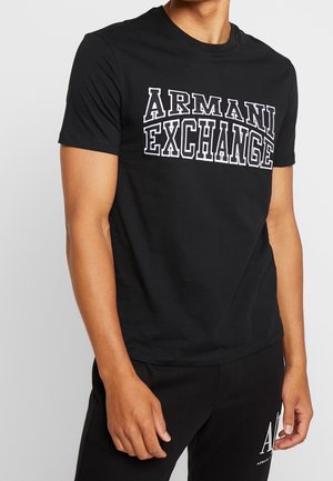 Homme portant un t-shirt noir Armani Exchange avec un logo blanc audacieux sur la poitrine et un pantalon noir assorti avec un logo blanc près de la poche.