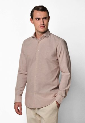 Uomo con camicia rosa chiaro a bottoni e pantaloni beige, che guarda leggermente verso sinistra, con le mani rilassate lungo i fianchi.