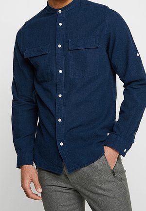 Mand iført en mørkeblå skjorte med knapper med to brystlommer og grå bukser med lommer med lynlås, den ene hånd i lommen.