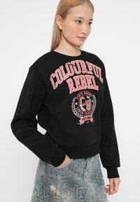 Zwarte sweatshirt met 'Colourful Rebel'-graphic in roze. Heeft een ronde halslijn en brede manchetten. Gecombineerd met gemusterde lichtblauwe jeans.