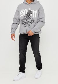 Felpa grigia con zip e stampa grafica, abbinata a jeans neri e sneaker bianchi. La felpa presenta un design ampio e un tessuto morbido.
