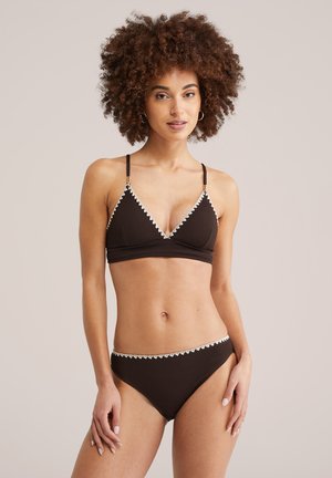 Ženska s kodrastimi lasmi, oblečena v črni bikini z belim robom, stoji pred enobarvnim bež ozadjem in gleda proti kameri.