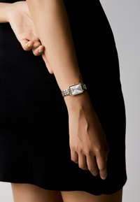 Reloj de cara cuadrada con eslabones de metal plateados y dorados, esfera blanca y marcadores de hora negros, llevado en una muñeca contra un fondo negro.