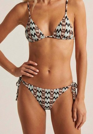 Mujer vistiendo un bikini triangular con estampado de rayas en zigzag negras, blancas y marrones, de pie con una mano en la cadera sobre un fondo neutro.