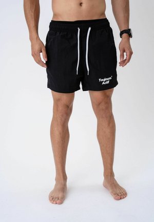 Mann trägt schwarze Sportshorts mit weißen Kordeln und dem Schriftzug „Toujours Actif“ am linken Bein, steht barfuß vor weißem Hintergrund.