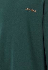 Mörkgrön sweatshirt i bomull med rund halsringning och en liten orange texttryck "LES'ARCS" på bröstet. Slät textur.