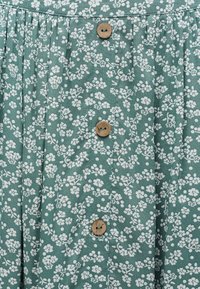 Grønt stof med lille hvidt blomster mønster, med lodrette folder og en række med fire runde træknapper.