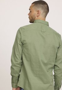 Chemise à manches longues en vert olive, en tissu texturé. Dispose d'un col classique, de poignets à boutons et d'un discret détail de parement au dos.