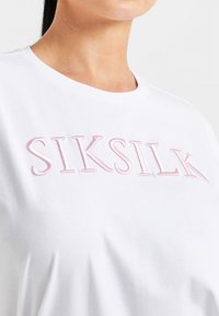 Bílý bavlněný tričko s růžovou vyšívanou logem "SIKSILK" vpředu, s kulatým výstřihem a krátkými rukávy. Hladká textura.