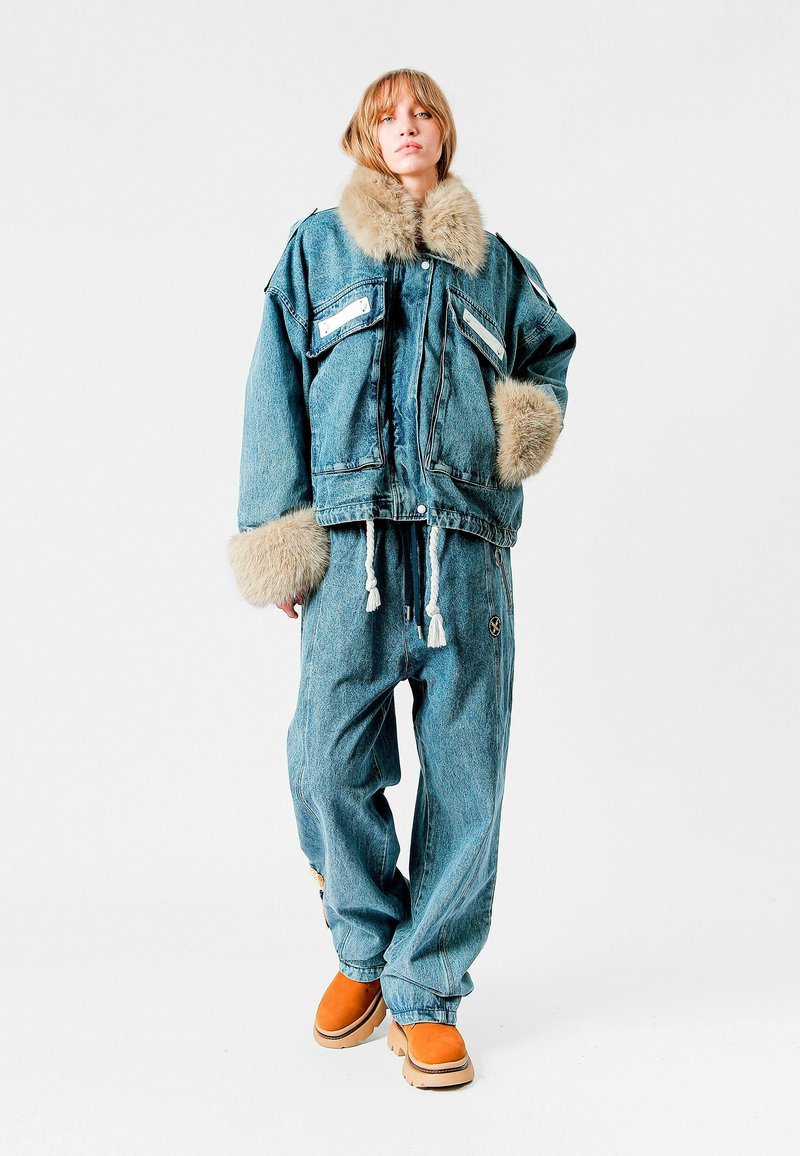 Chaqueta de denim con cuello de pelaje, dos bolsillos en el pecho. Combinada con pantalones de denim de corte holgado. El modelo lleva botas naranjas y está de pie sobre un fondo blanco.